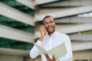 Black man in a white shirt holds a folder in his left hand and talks on his phone with his right hand. He is standing in front of a large building with a modern design photo