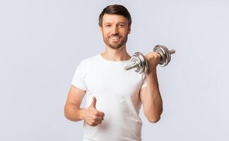 A man in a white t-shirt is lifting a dumbbell with his right arm and giving a thumbs up with his left hand. He is smiling and looks happy and healthy. photo