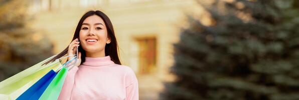 Asian woman is standing outdoors holding a shopping bag in one hand while talking on a cell phone. She appears focused on her conversation, with a determined expression on her face, web-banner photo