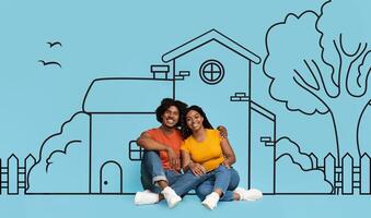 A cheerful young black couple is sitting closely together against a backdrop with a drawing of a house, trees, and birds. The drawing implies dream or goal of homeownership, or simple life together photo
