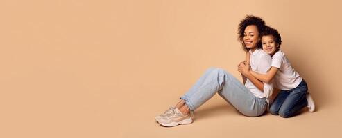 African American mother and her son share a joyful moment, sitting closely on a light-colored backdrop. They are both wearing casual, comfortable clothing and radiating happiness. photo