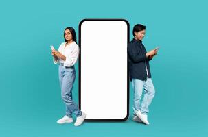 Multiracial couple stand back to back, each absorbed in their own smartphone, with a large, vertical, blank white screen between them on a turquoise background photo