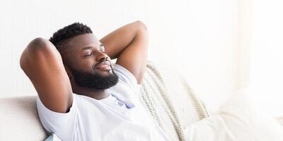 A young African American man with a beard is sitting on a white couch at home. He is leaning back with his arms behind his head and his eyes closed, appearing relaxed and content, copy space photo