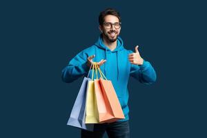 un hombre vistiendo un azul capucha es en pie mientras participación compras pantalones en su manos. él es sonriente y dando un pulgares arriba gesto, expresando satisfacción con su compras foto