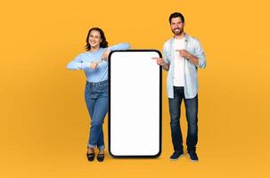 A man and woman are standing in front of a blank screen, both pointing towards it with their index fingers. The background is a solid yellow color, creating a vivid contrast with the neutral screen. photo