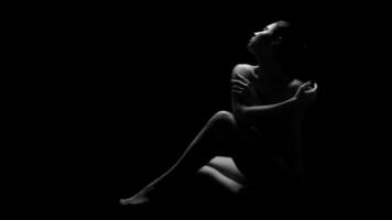A black and white photograph of a woman sitting in darkness, illuminated only by a single light source. She has her arms crossed, and her gaze is directed upwards. photo