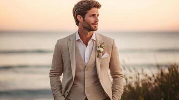A groom stands confidently by the shoreline during sunset, dressed in a tailored suit with a floral accent, as ocean waves softly lap at the shore. photo