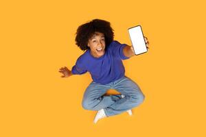 A young man with curly hair sits on a yellow background, holding a smartphone with a white screen towards the camera. He is smiling and looking at the viewer, top view photo