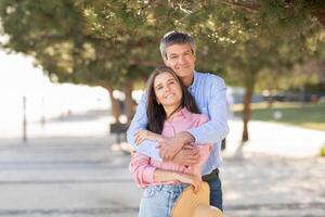 A happy couple stands close together on a beach promenade, surrounded by trees, embracing each other with smiles. The sun shines brightly, creating a warm atmosphere during their outing. photo