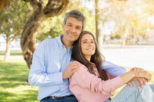 A man and woman relax together outdoors, sitting on the grass beneath a large tree as sunlight filters through the leaves, creating a warm and inviting atmosphere. photo