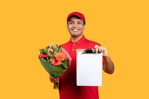 A smiling delivery man, wearing a red uniform and cap, holds a beautiful flower bouquet in one hand and a clipboard in the other, standing against a vibrant yellow background. photo