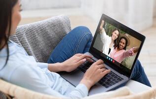 A woman is sitting on a couch at home, using a laptop to chat with two friends. The friends are smiling and waving at the camera, while the woman is typing on the keyboard. photo