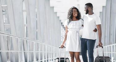 A young black couple, dressed in casual attire, walks arm-in-arm through a modern airport terminal. They are both smiling and appear to be happy and relaxed, as they head towards their destination photo