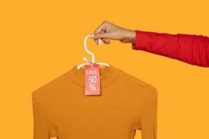 This image shows a hand holding a yellow mock-up sweater on a hanger. The sweater has 50 percent off sale tag attached to it. The background is a bright yellow color, cropped photo