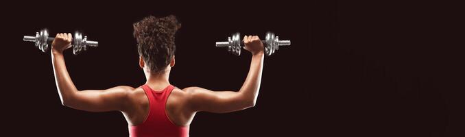 Back view of african bodybuilder woman working out with dumbbells over black studio background, panorama photo