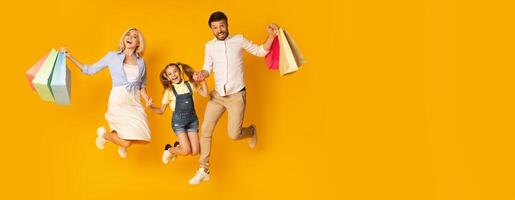 A cheerful family of three is leaping in the air while holding colorful shopping bags. They display bright smiles, highlighting their excitement. The backdrop is striking yellow, copy space photo