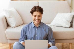 A man sits on the floor in front of a couch, smiling as he uses a laptop. photo