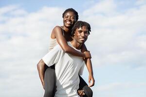 Happy black couple enjoying a piggyback ride on a sunny day. The woman is sitting on the mans back, and they are both smiling and looking at the camera. photo