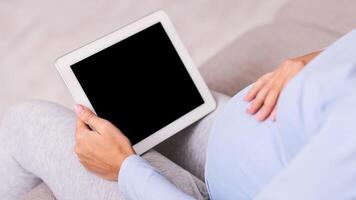 Pregnancy App. Unrecognizable Expectant Lady Using Digital Tablet With Empty Screen Sitting On Couch Indoor. Mockup, Cropped photo