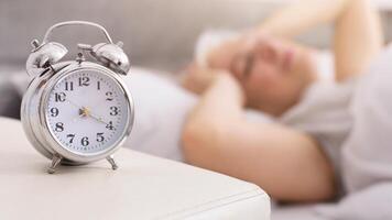 An alarm clock with a digital display sits atop a clean white table, casting a shadow in a well-lit room. The clocks numbers indicate the current time over sleeping senior lady photo