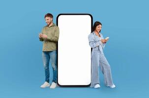 A man and woman are positioned beside a blank screen on a blue background. Both individuals are looking towards the screen in a professional setting. photo