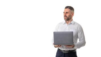 A professional businessman, dressed in a white shirt and dark pants, stands confidently holding a laptop in a bright office environment with a clean, white background, copy space photo