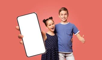 Two children, a boy and a girl, stand in front of a pink background. The boy is giving a thumbs up. The girl is holding a smartphone with a blank white screen. photo