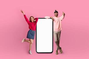 Indian man and european woman are energetically posing with smiles, standing beside an oversized model of a smartphone with a blank screen photo