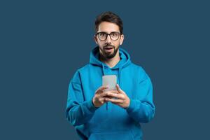 A man wearing a blue hoodie is standing while holding a cell phone in his hand, looking at the screen. He seems focused on the device, possibly texting or browsing the internet. photo