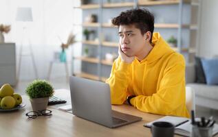 Bored young Asian guy sitting at desk using pc laptop, looking aside, thinking about something during online lesson or remote work at home office. Tired man having dull distant job, feeling lazy photo