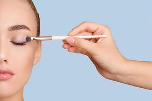 This image shows a womans face with her eyes closed as she applies eye shadow with a brush held by another hand. The womans skin is clear, and the image is against a light blue background. photo