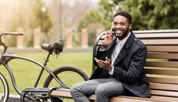 Black business professional is using his phone in a park, seated on a bench with a bicycle beside him. The scene is calm and serene, highlighting a balance between work and nature. photo