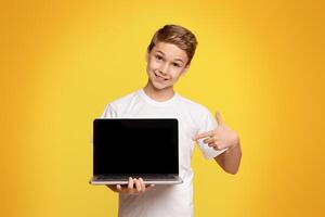 Smiling teenager boy pointing at blank screen of laptop, recommend educational website or application, mockup photo