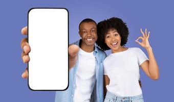 A young African American couple stands together in front of a purple background, smiling and looking at the camera. The man is holding a smartphone with a blank screen, woman showing okay photo