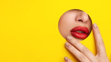 A close-up view of a woman lips peeking out of a hole against a vibrant yellow background. The lips are painted in a bold red color, creating a striking contrast with the cheerful yellow hue. photo