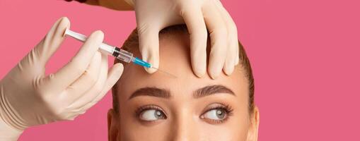 A woman is receiving a forehead injection with a syringe. A gloved hand is holding the syringe, and another gloved hand is holding the woman forehead, web-banner photo