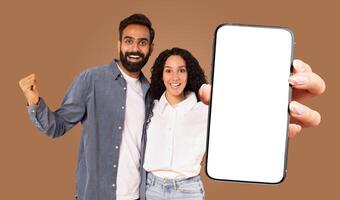 A happy multiracial couple is standing in front of a brown background, holding a large smartphone with a blank screen. photo