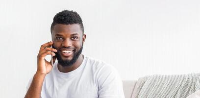 A young Black man with a beard is smiling while talking on his phone. He is sitting on a couch wearing a white t-shirt. The background is a white wall, copy space photo