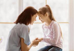 A mother and daughter are shown in a close-up shot, seated by a window, smiling at each other and holding hands. The image portrays a sense of love, connection, and warmth. photo
