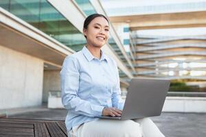 Asian woman is sitting on a wooden bench outdoors, using a laptop. She appears focused on the screen, with one hand typing on the keyboard. The background shows a park setting with trees. photo