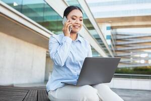 Asian woman is seated on a bench, engrossed in a phone conversation while simultaneously working on a laptop. She appears focused and multitasking efficiently in a public setting. photo