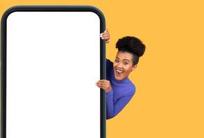 A young woman with curly hair joyfully leans out from behind a large mobile device, showcasing her excitement. The bright yellow backdrop adds a cheerful touch, enhancing the playful atmosphere photo