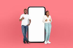 On a deep pink background, a black couple, both smiling, stands adjacent to an enormous smartphone with a blank display, ready for showcasing apps or designs. photo