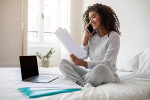 Hispanic young woman, comfortably dressed in casual athleisure wear, sits cross-legged on a white bedspread. Engaged in a phone conversation, she holds a document in one hand with a laptop open photo