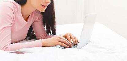 A woman is lying on a bed, focused on using a laptop computer. She is typing and browsing the internet while comfortably positioned. The room is softly lit, cropped photo