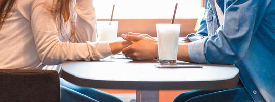 Two individuals, seated at a wooden table, are holding hands in a display of closeness and connection. The gesture suggests intimacy and mutual affection between the pair, cropped photo