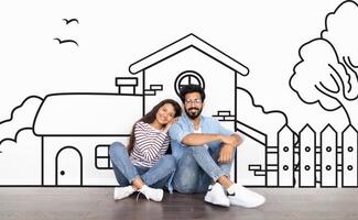 Cheerful young indian couple sitting by white wall with a house outline, dreaming of a home. Real estate, mortgage concept photo