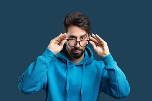 un enfocado hombre con un barba y despeinado pelo es ajustando su lentes y mirando Derecho adelante. él es vistiendo un vibrante azul sudadera, cuales contrastes con el sólido azul fondo foto