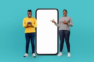 Two smiling black people interact with their mobile devices while standing on a turquoise background. A large, blank phone frame stands between them as one person points at it photo