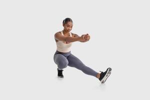 African American woman is shown doing a squat exercise on a plain white background. She is bending her knees and lowering her hips while keeping her back straight and arms extended in front of her photo